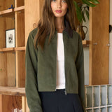 Woman wearing a green jacket and black skirt standing in a room with wooden shelves and a plant.