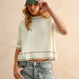 Person wearing a light-colored sweater with striped sleeves and a cap, standing against a beige background