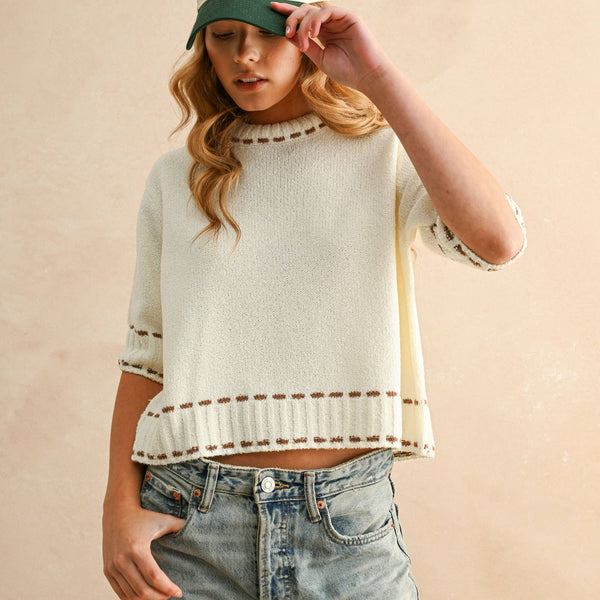 Person wearing a light-colored sweater with striped sleeves and a cap, standing against a beige background