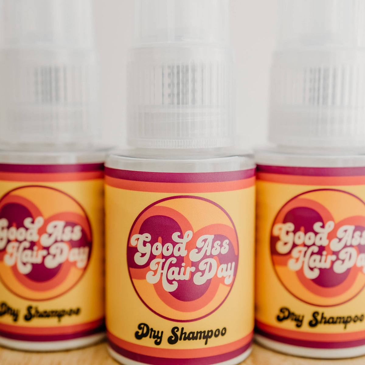 Three bottles of 'Good As Hair Day' dry shampoo on a wooden surface.