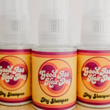 Three bottles of 'Good As Hair Day' dry shampoo on a wooden surface.