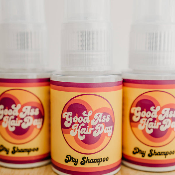 Three bottles of 'Good As Hair Day' dry shampoo on a wooden surface.