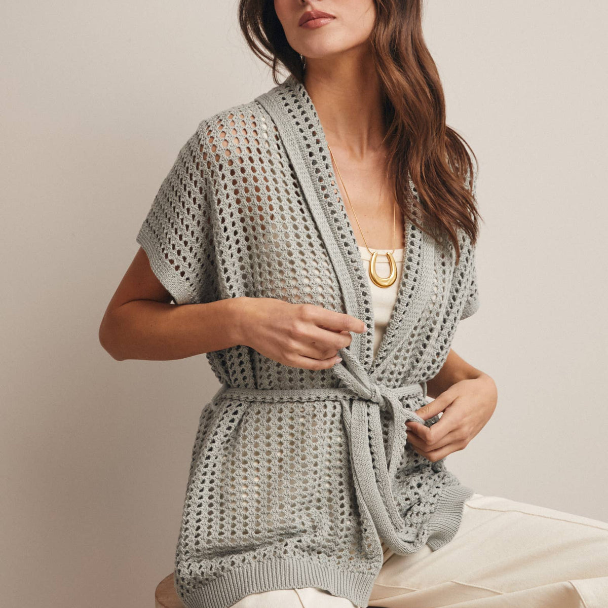 Woman wearing a Sage knit Cardigan with open knit and waist tie sitting on a wooden stool against a plain background