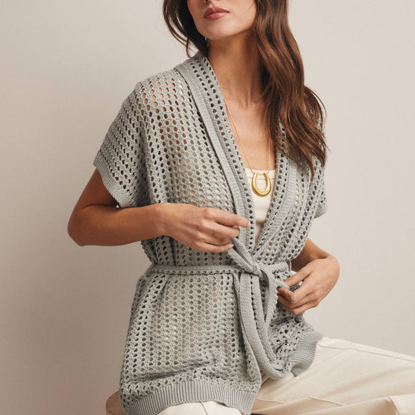 Woman wearing a Sage knit Cardigan with open knit and waist tie sitting on a wooden stool against a plain background